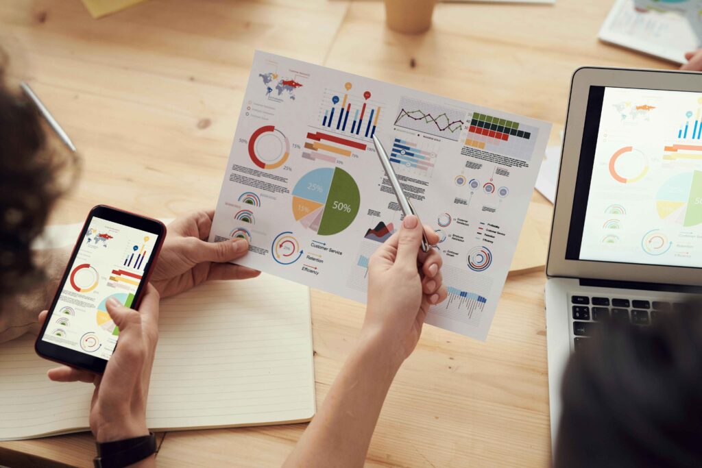 People analyzing colorful business charts and graphs on paper, a smartphone, and a laptop during a meeting at a wooden desk.